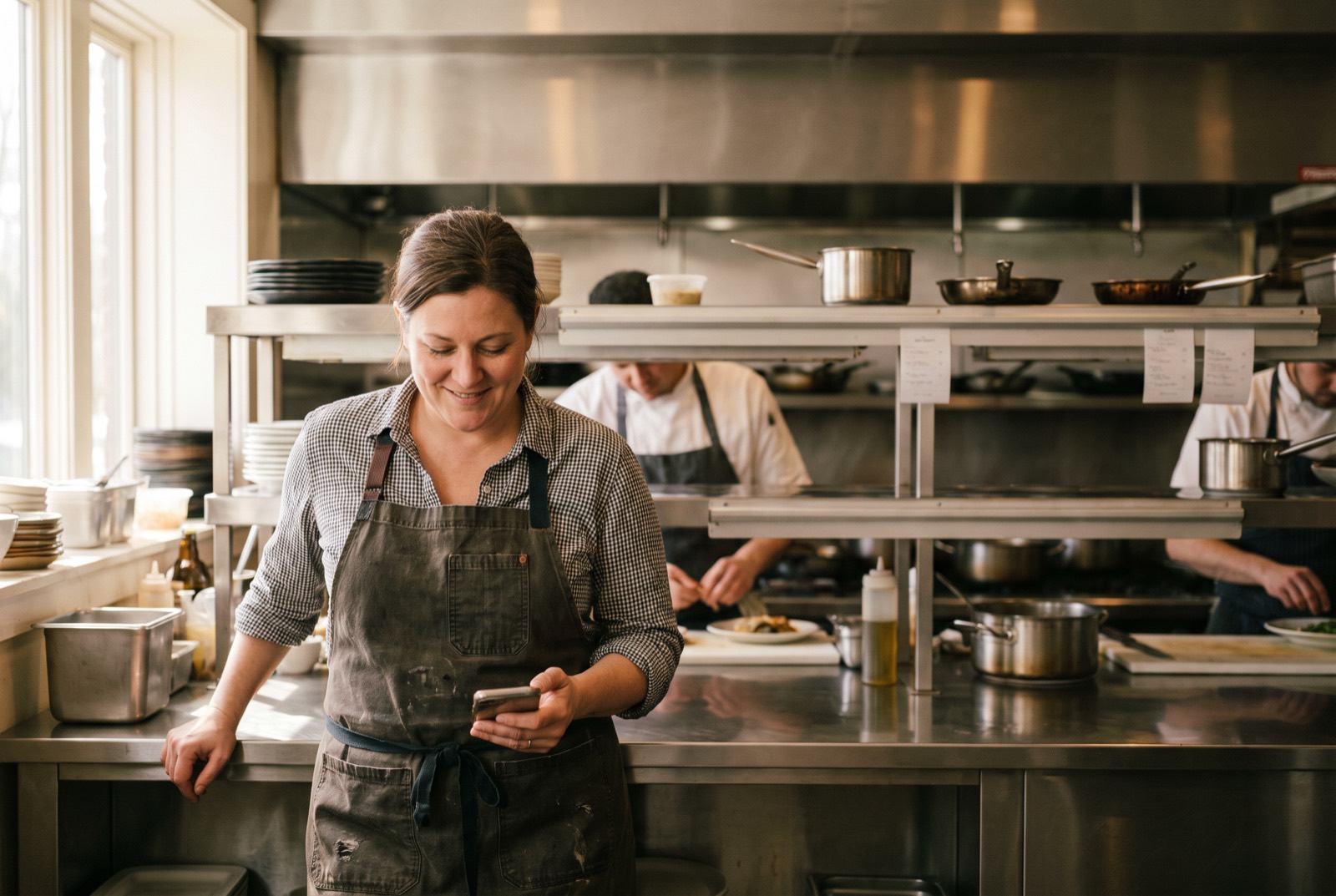 A restaurant owner in an apron reads a compliance record on her phone at the pass in her own kitchen.